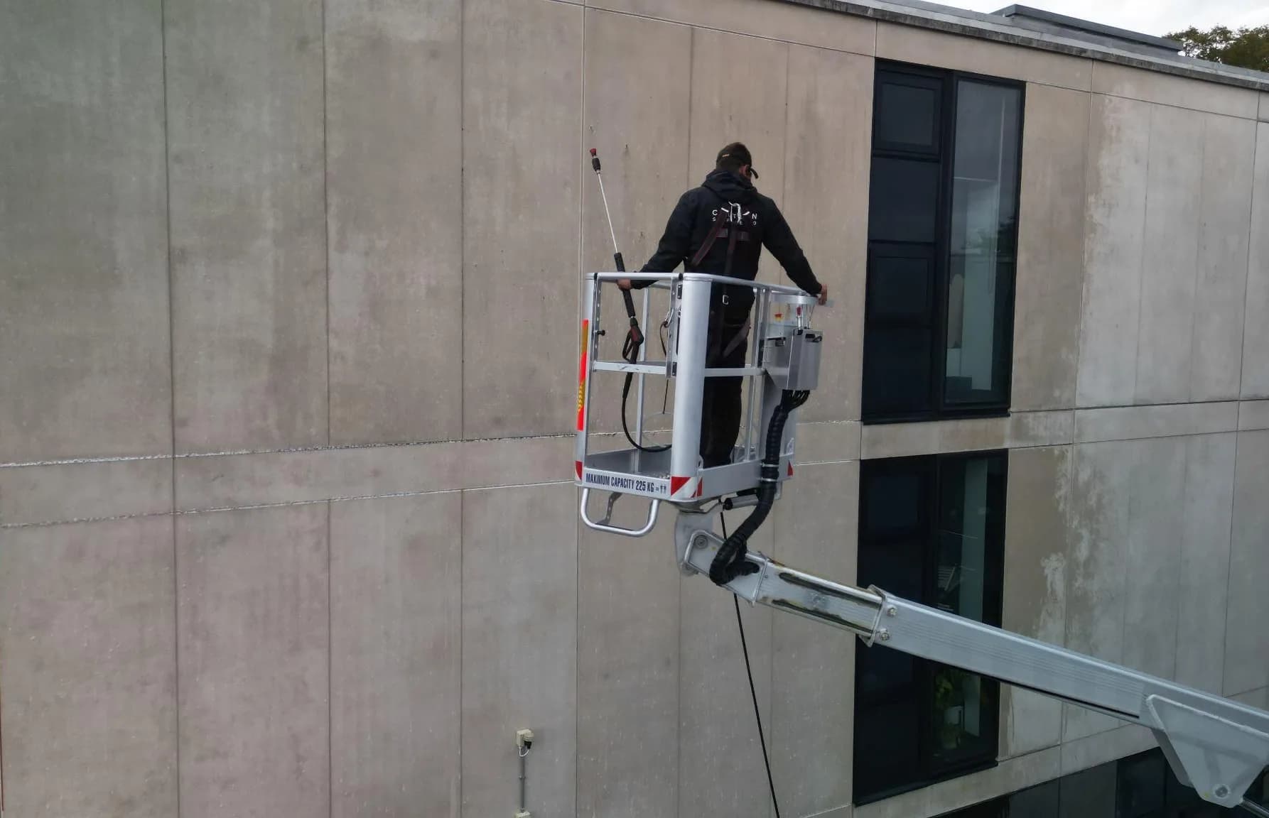 Harnessed technician in a lift basket washing large stained stone facade panels, seen from behind