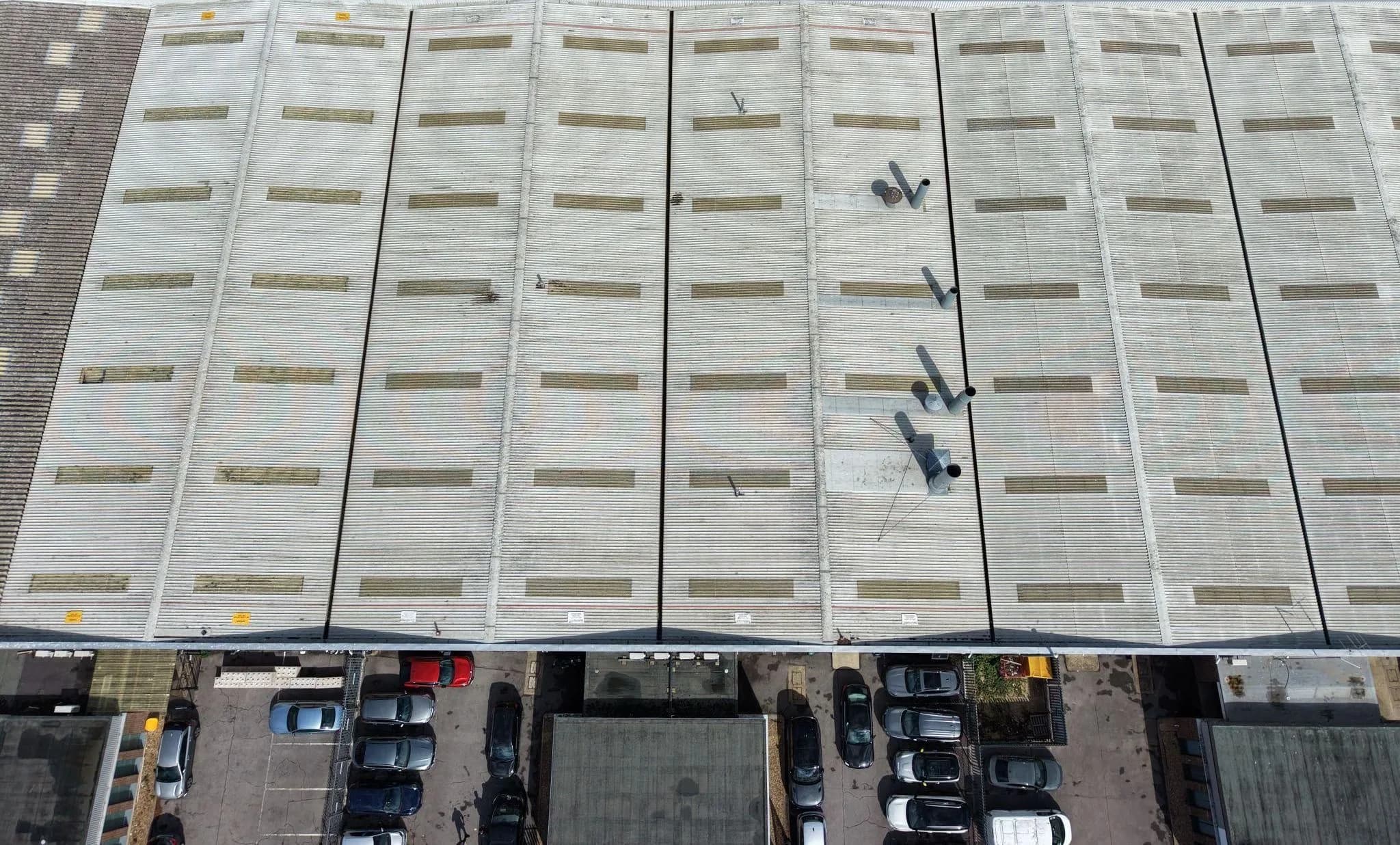 Overhead drone view of a long corrugated factory roof with rows of rooflights beside a car park