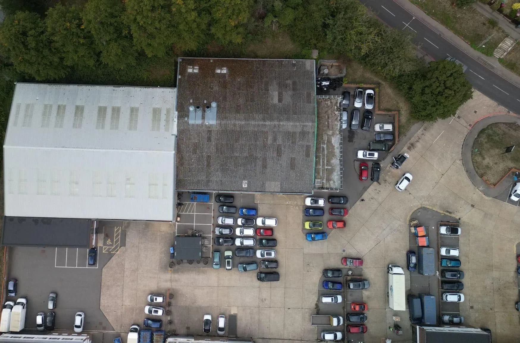 Overhead drone view of two industrial units, their roofs, a concrete service yard and a busy car park