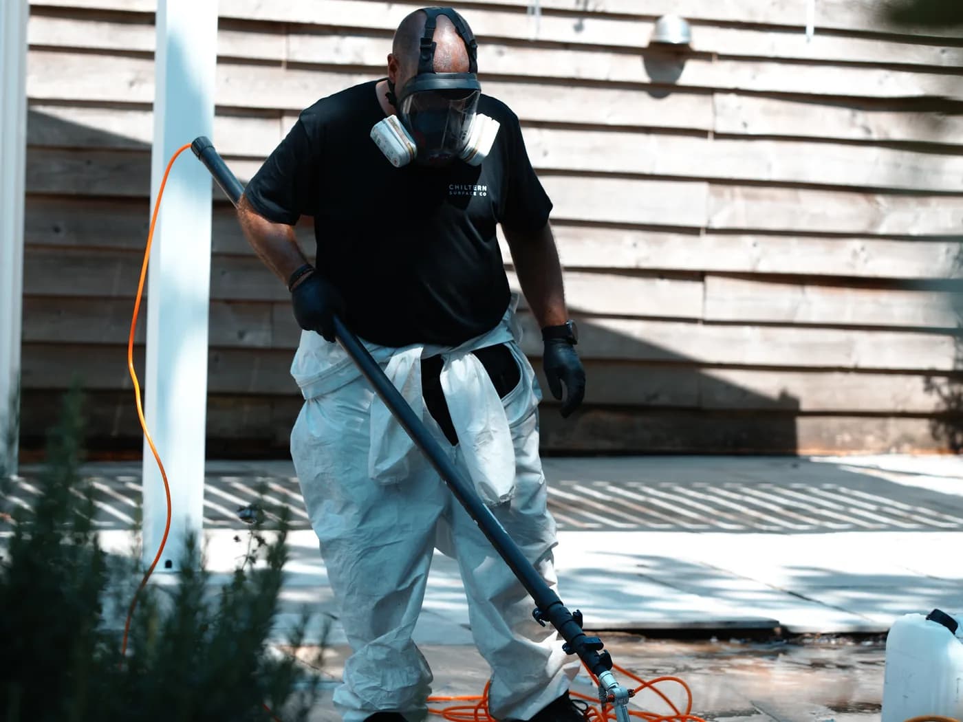 Chiltern EverGuard™ being applied over the cleaned sandstone from a long spray lance fed by a tank standing on the paving, with the timber fence alongside