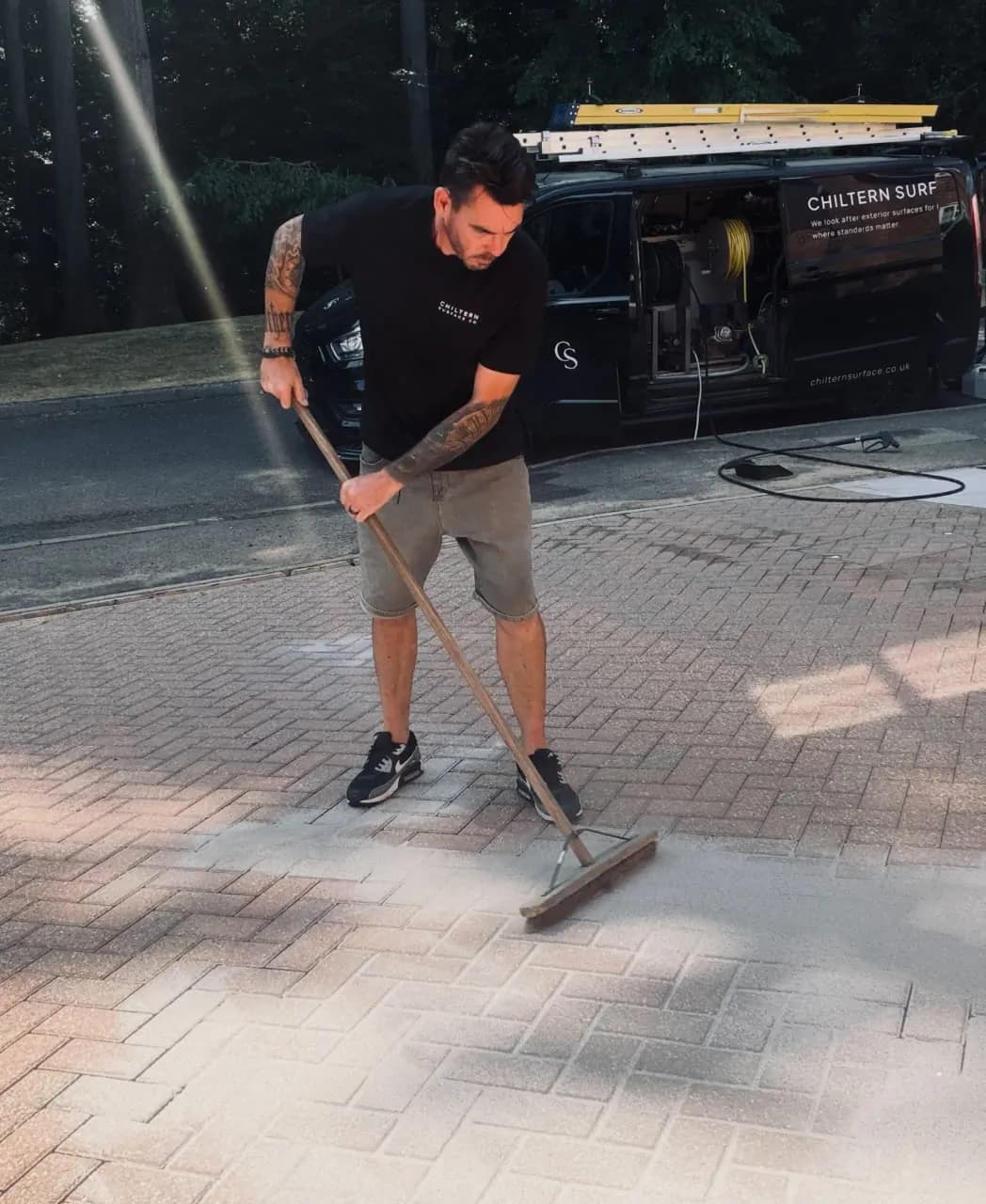 Operative brushing jointing sand into herringbone block paving, a work van with its side door open behind him
