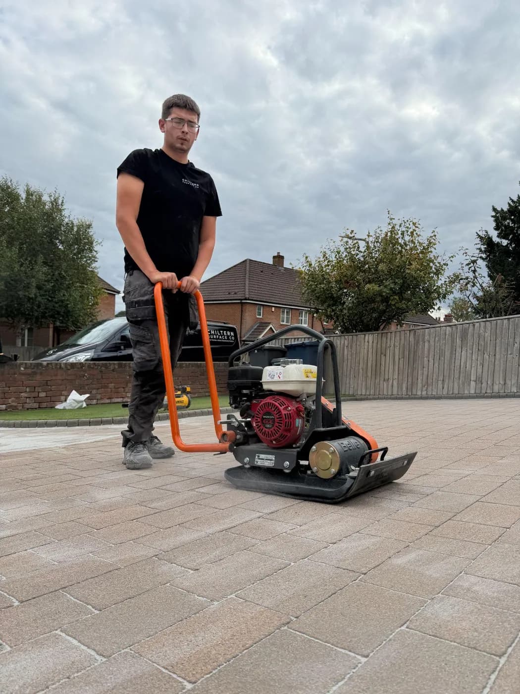 Operative guiding a plate compactor over freshly sanded block paving, red-brick houses and a timber fence behind