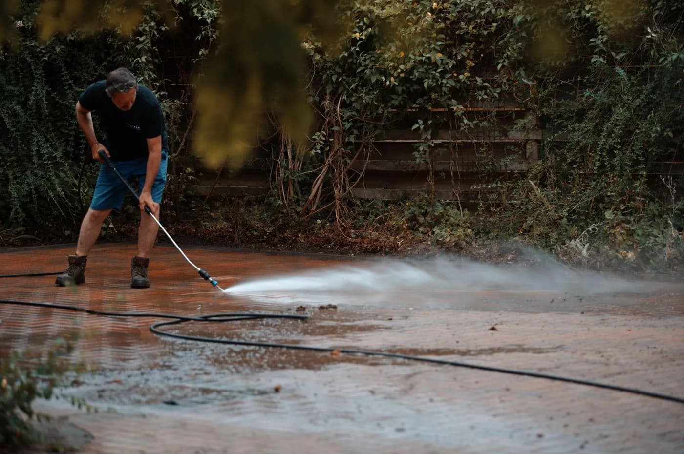Operative cleaning red herringbone block paving with a lance, spray lifting dirt beside an ivy-covered fence