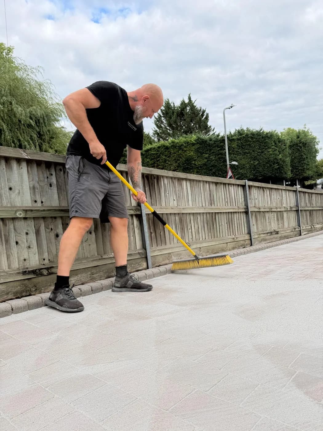 Operative sweeping jointing sand across pale block paving beside a timber fence, with tall hedging behind