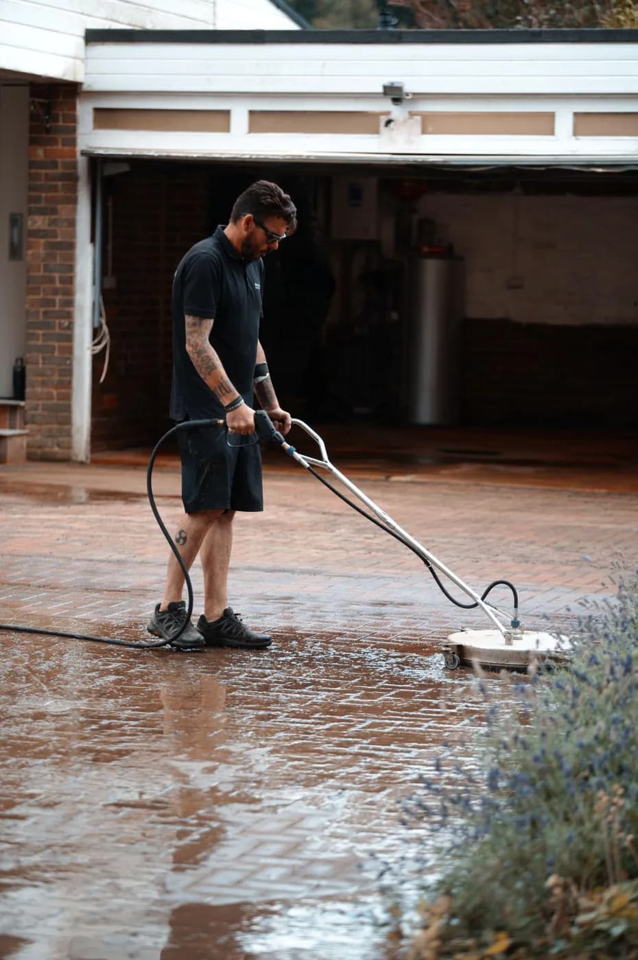 Operative running a rotary surface cleaner over wet red block paving in front of an open double garage