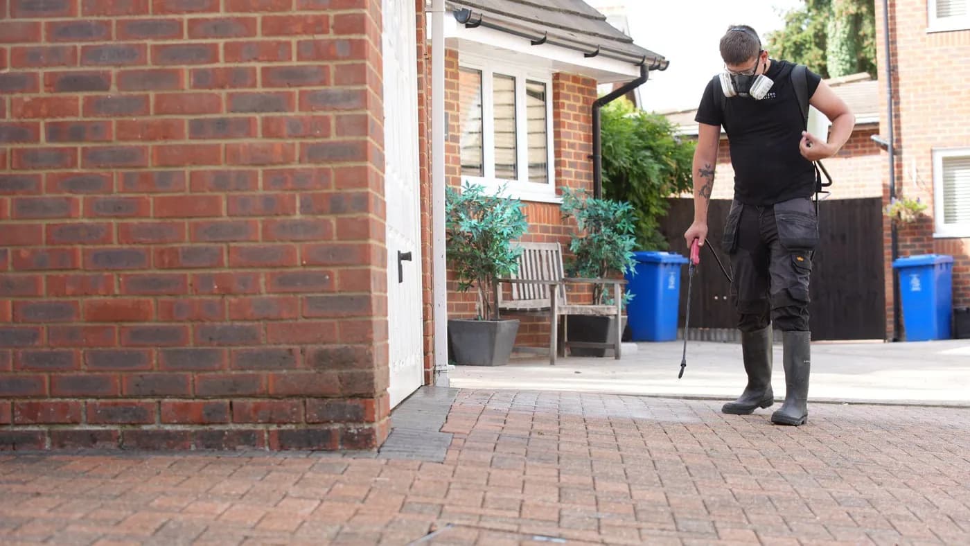 Operative in a respirator spraying treatment onto red block paving beside a brick garage, with blue bins behind