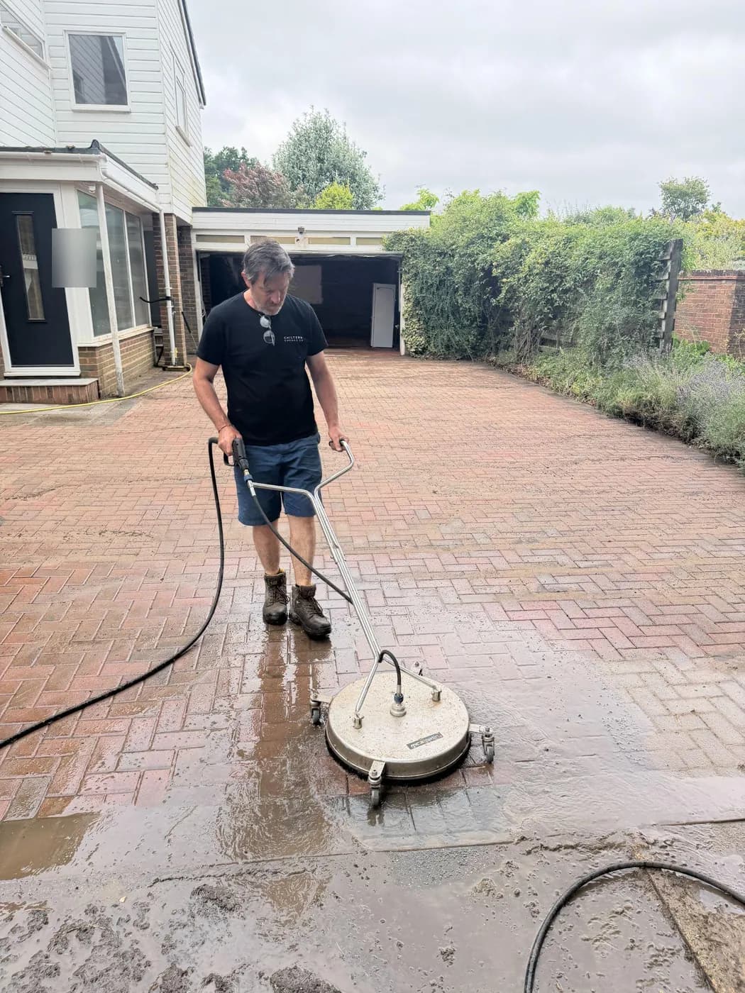 Operative running a rotary surface cleaner over red herringbone block paving, a clean strip showing against the dirty drive