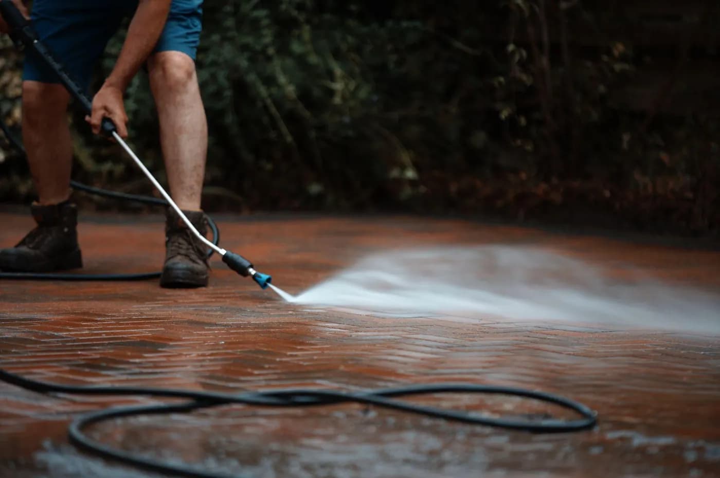 Technician in work boots rinsing wet red herringbone block paving with a lance, a coiled hose at his feet