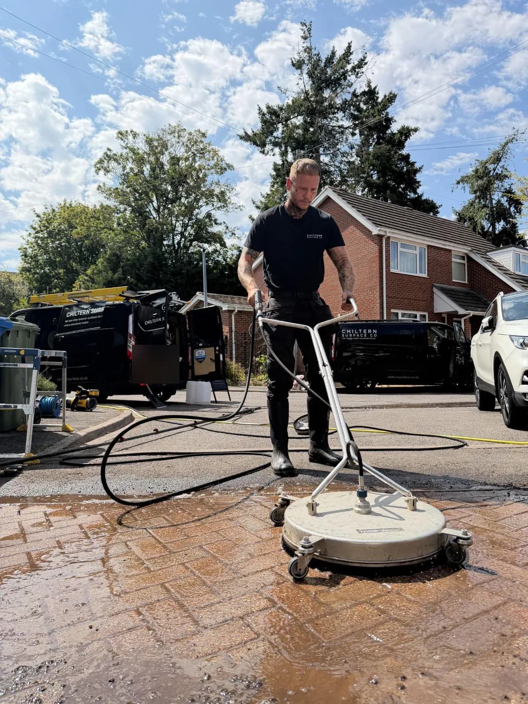 Operative pushing a rotary surface cleaner across wet block paving, two branded vans and brick houses behind