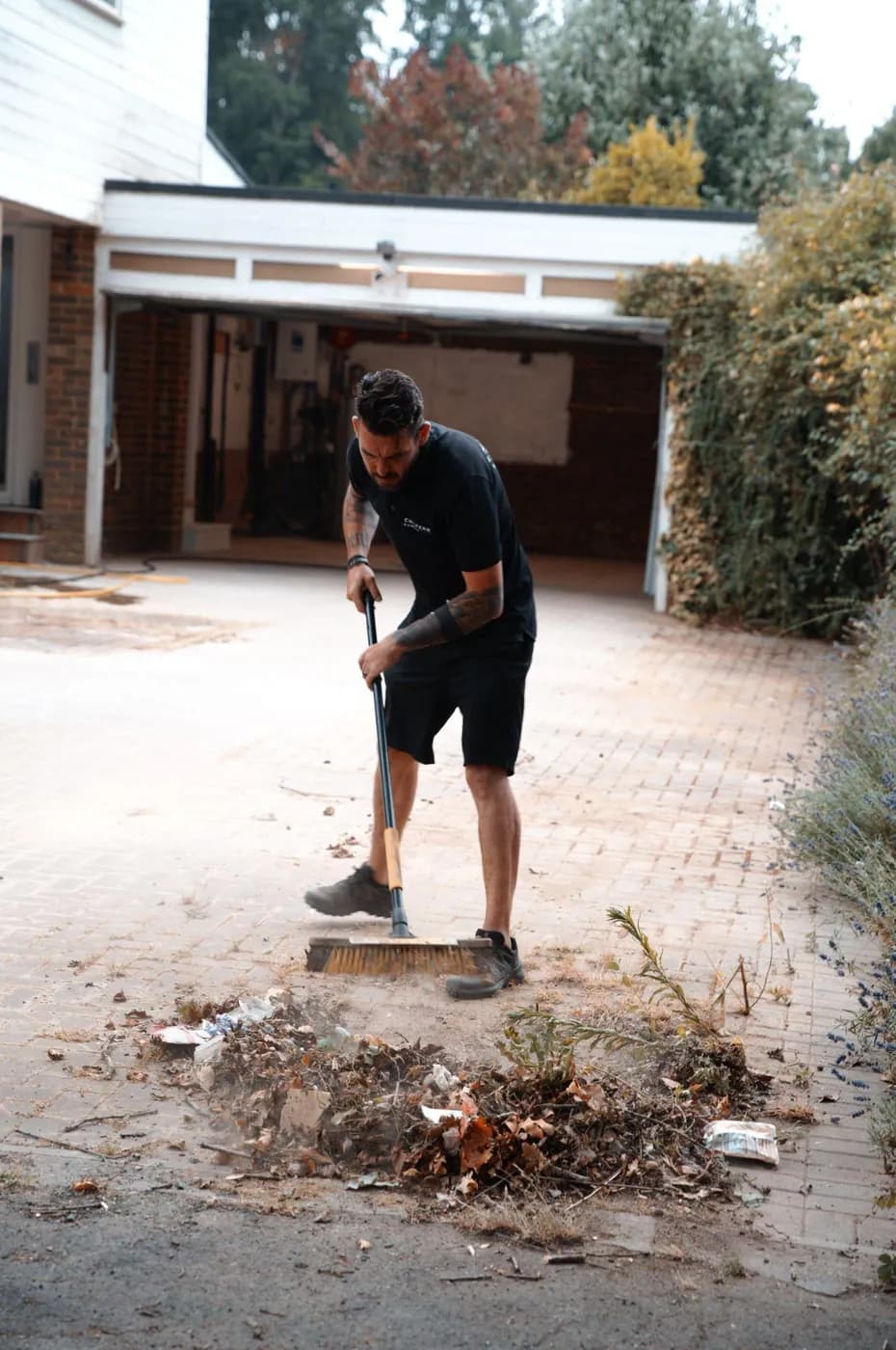 Operative sweeping fallen leaves, weeds and litter off a block-paved driveway before washing, an open garage behind