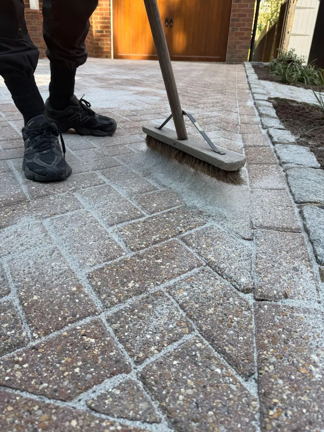 Broom sweeping jointing sand into herringbone block paving edged with granite setts, timber garage doors beyond