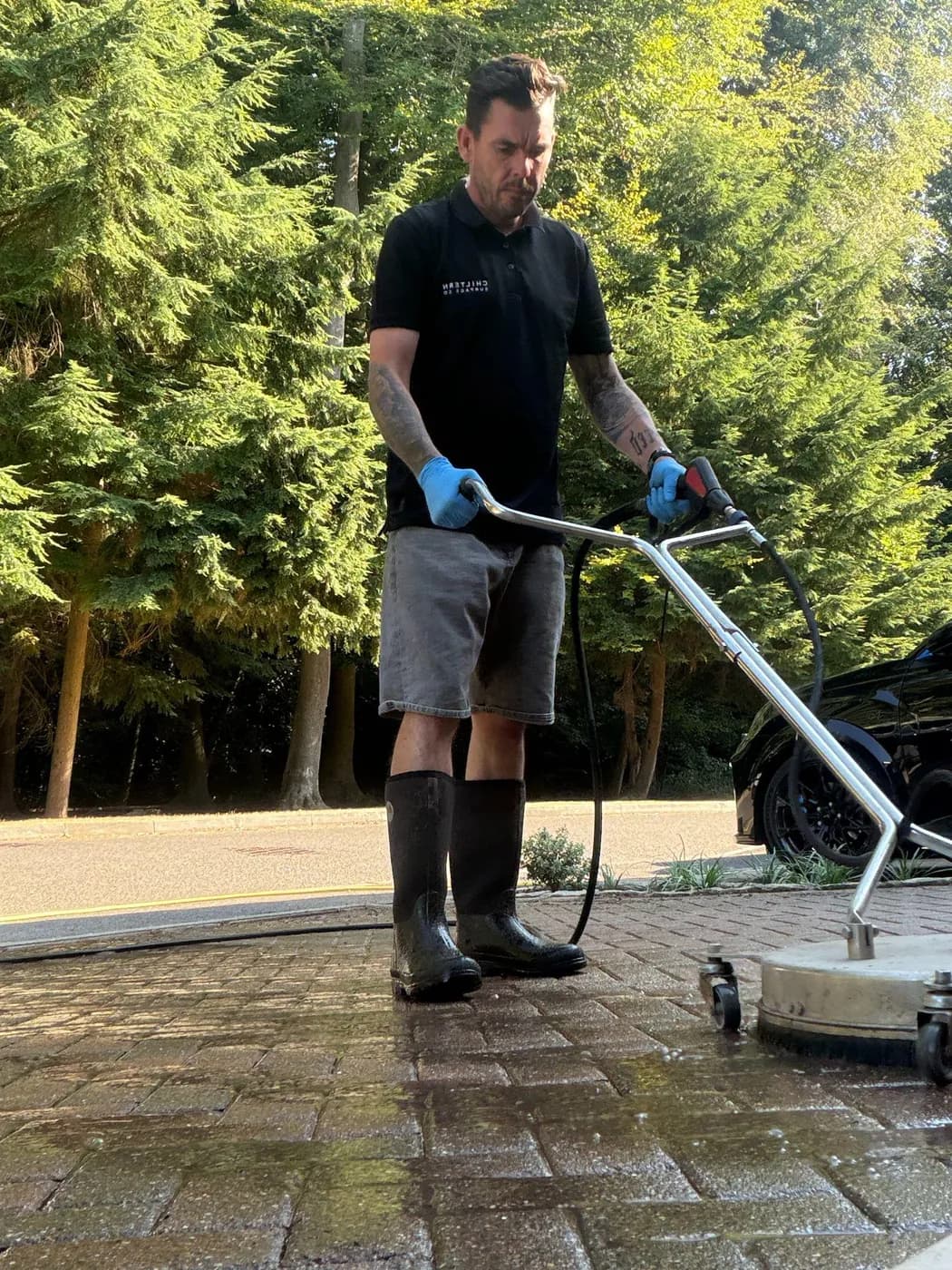Operative guiding a rotary surface cleaner across wet brindle block paving, with tall conifers and a parked car behind