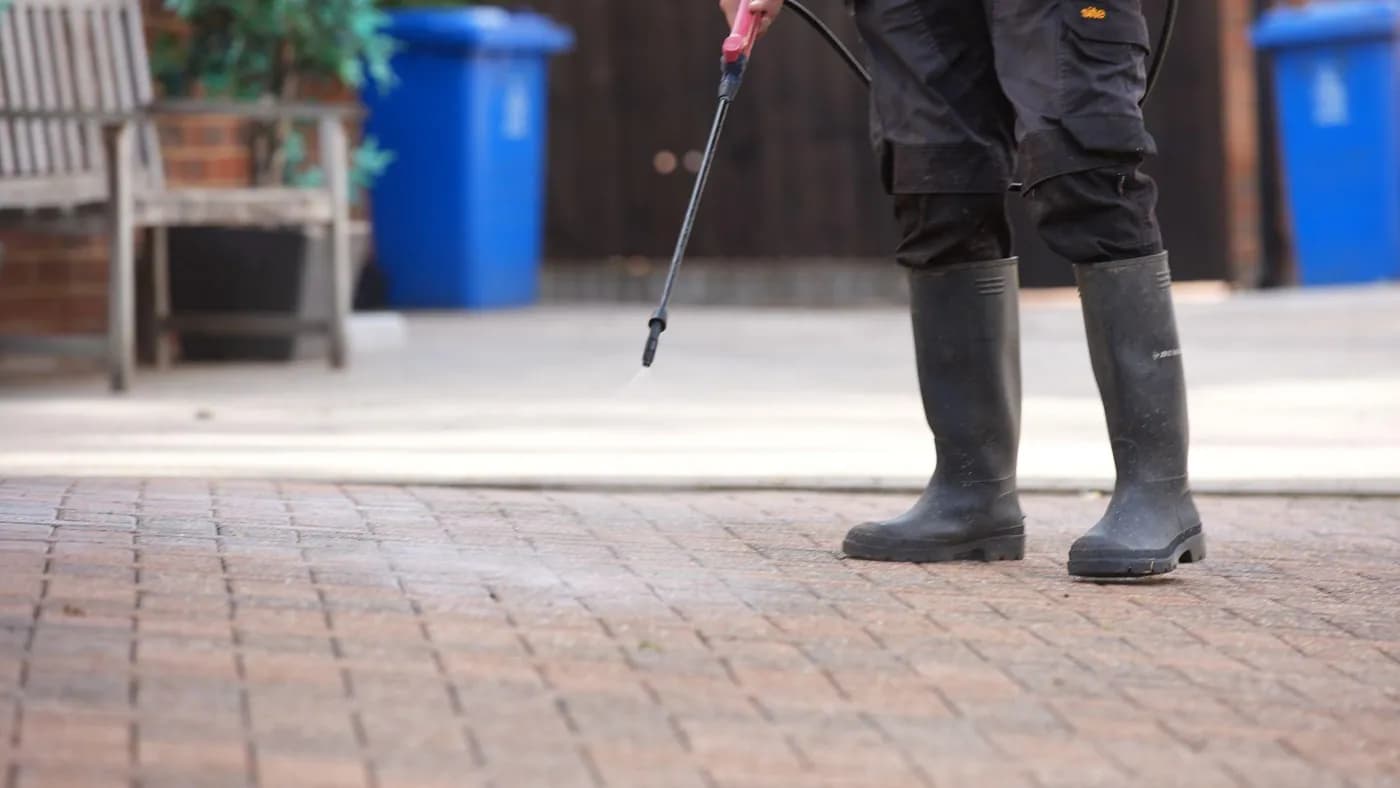 Close view of an operative's boots and sprayer lance misting treatment onto red block paving, a bench and bins behind