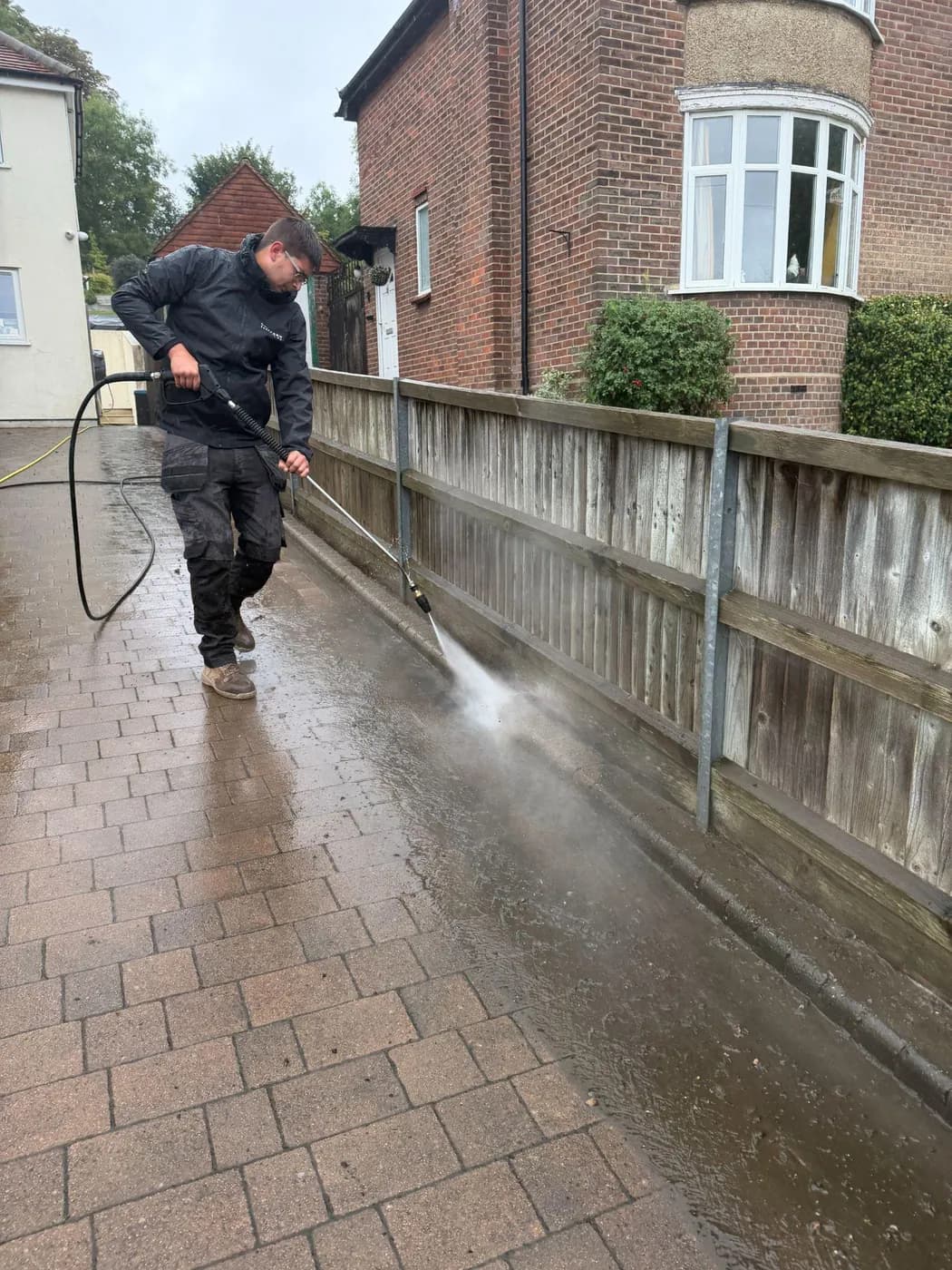 Operative rinsing the edge of a block-paved driveway with a lance along a timber fence, a bay-fronted brick house behind