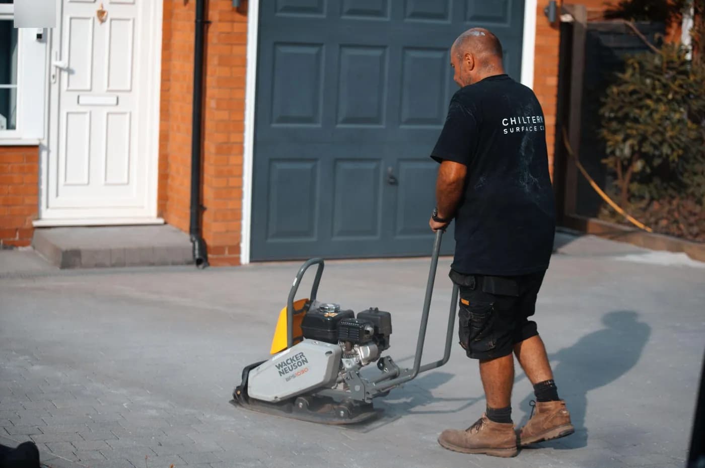 Operative pushing a plate compactor across freshly cleaned grey block paving in front of a garage