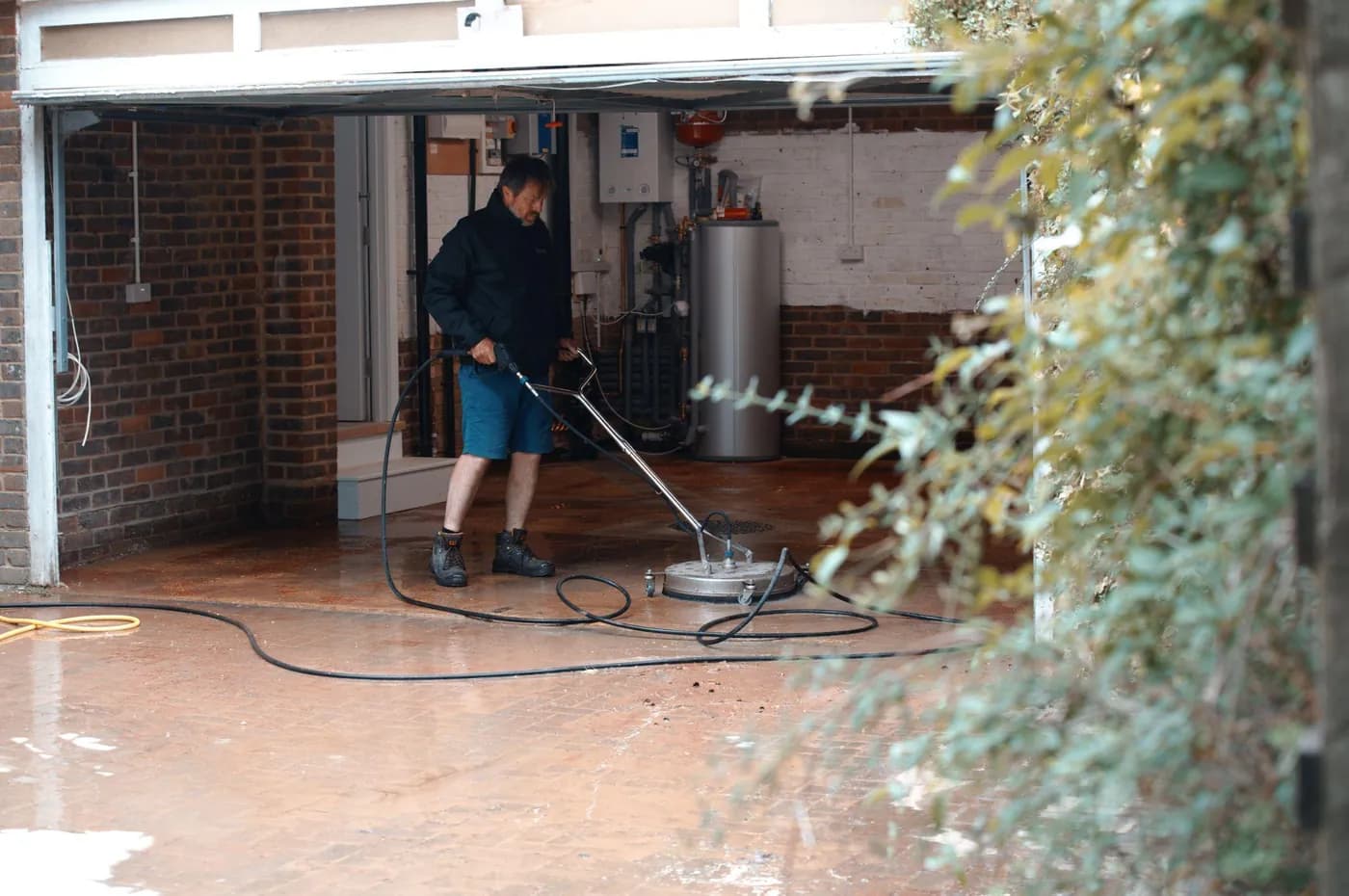 Operative guiding a rotary surface cleaner over wet block paving at the mouth of an open garage, foliage in the foreground