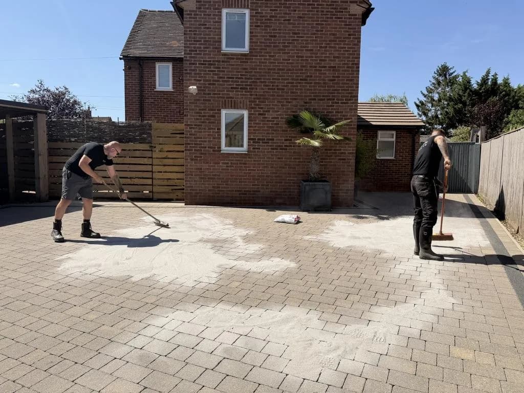 Two operatives brushing pale jointing sand across a cleaned block-paved driveway in front of a red-brick house