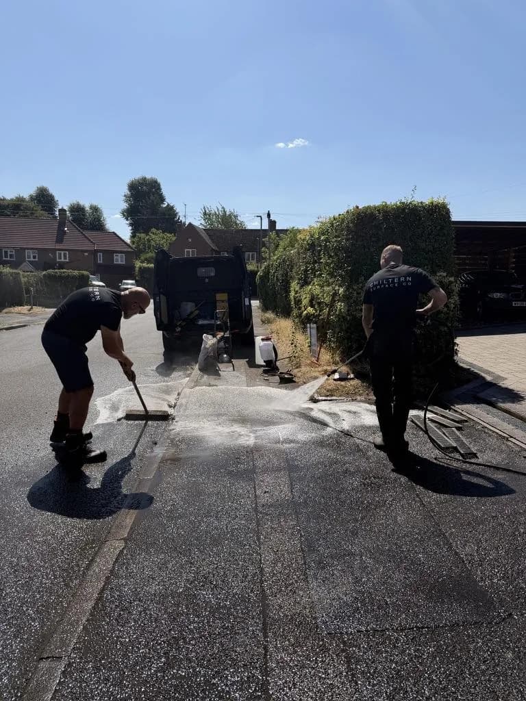 Two operatives rinsing and brushing a tarmac drive beside a channel drain and hedge, a van parked at the end