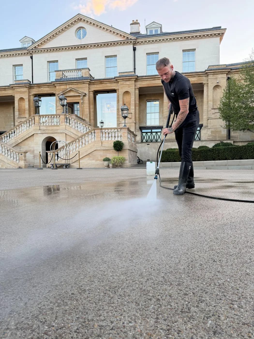 Operative cleaning a pale aggregate forecourt with a lance before a large classical stone house and twin staircase