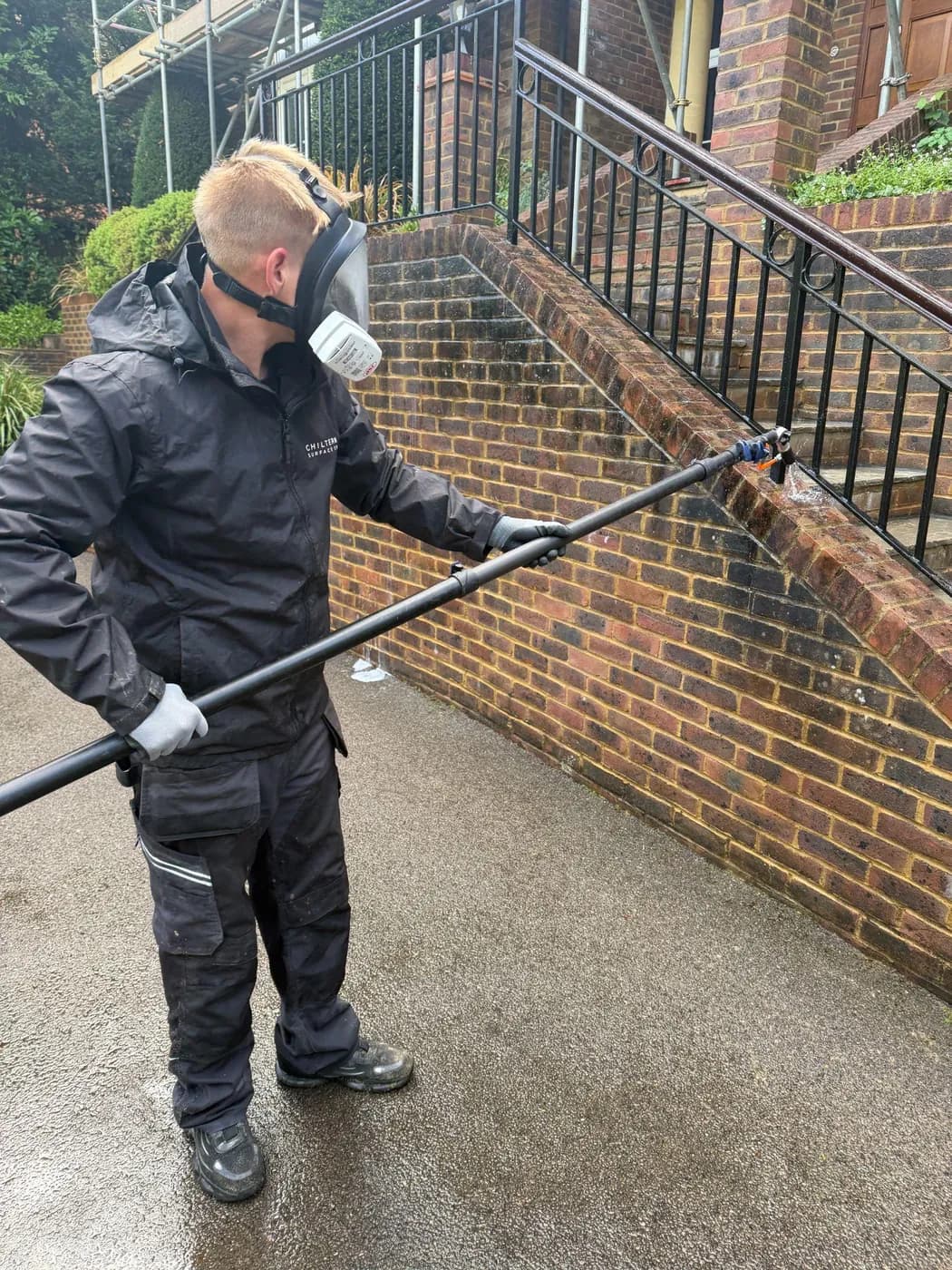 Operative in a full-face respirator applying solution from a long pole to a stained brick wall beside garden steps