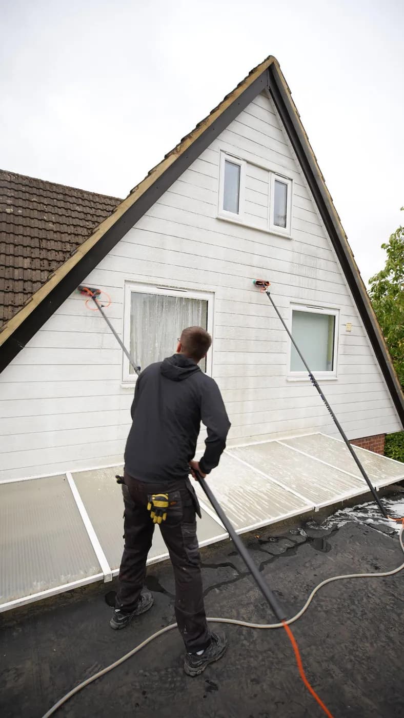 Operative on a flat roof soft washing a white clad gable with two water-fed brush poles, a lean-to roof beside him