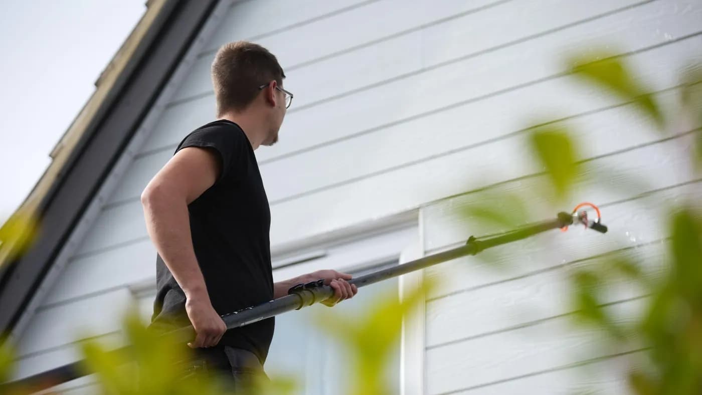 Operative using a long pole to apply a low-pressure spray to pale horizontal cladding on a gable, leaves blurred in front