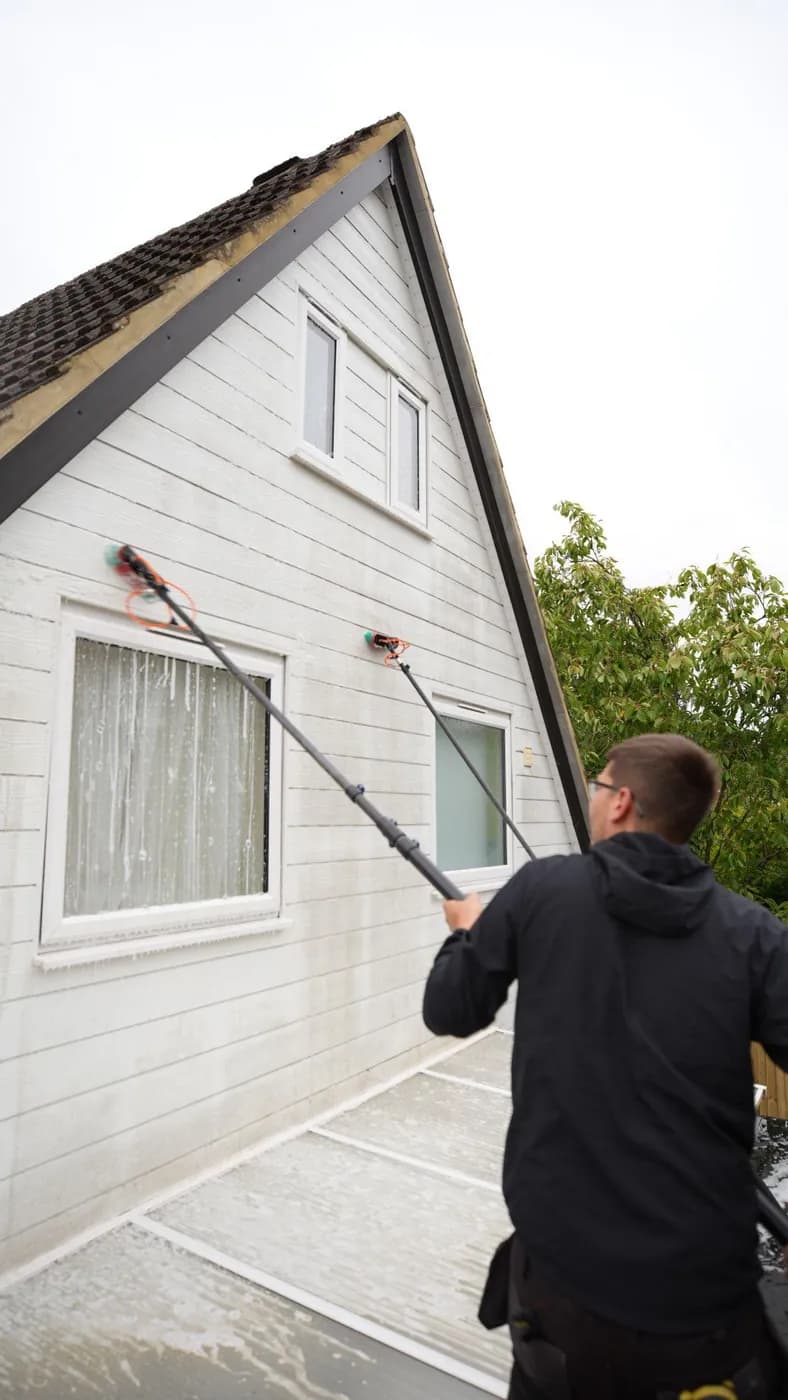 Operative using two water-fed brush poles to soft wash white cladding on a gable above a lean-to roof