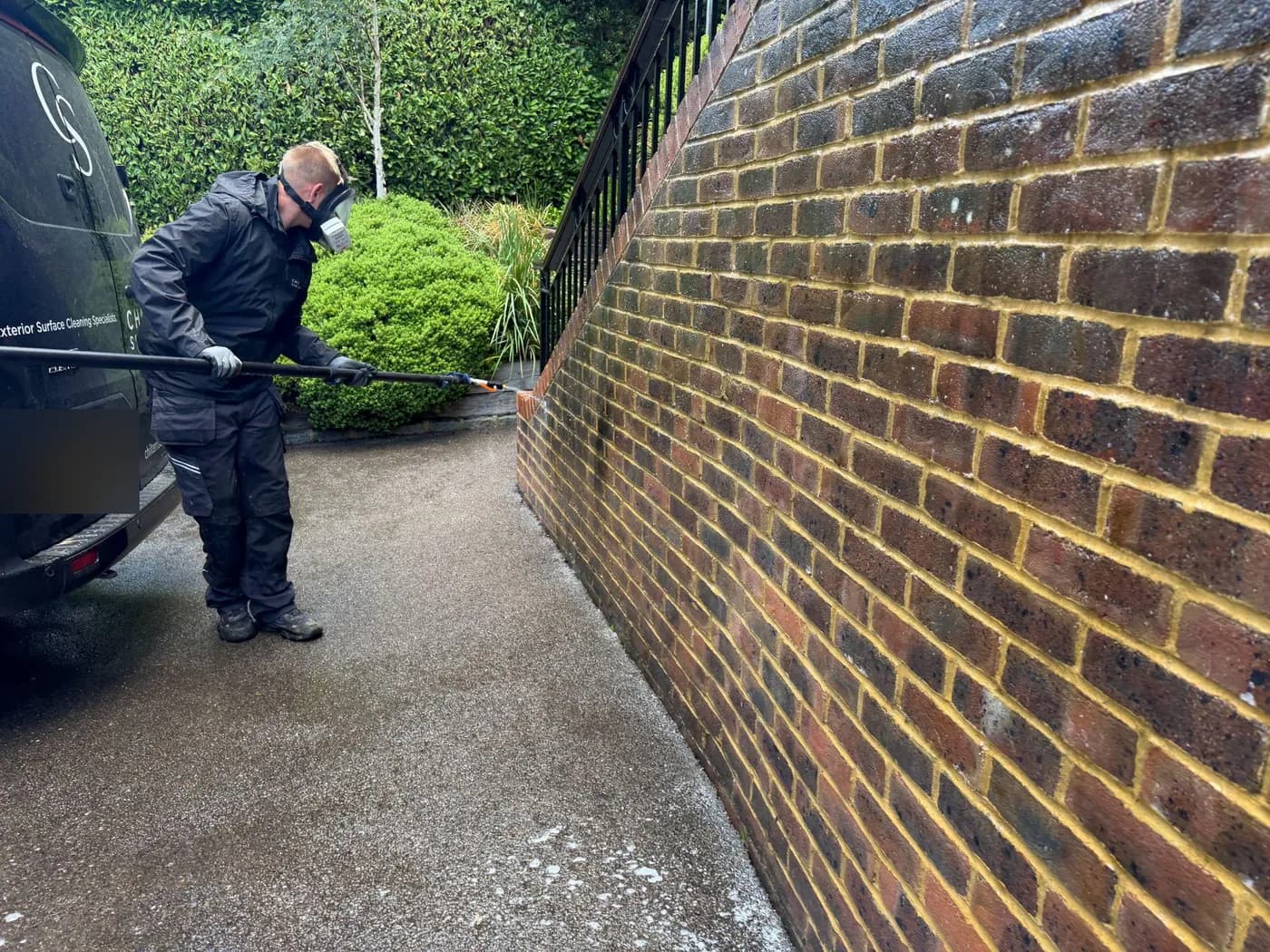 Operative in a respirator applying solution by pole to a brick retaining wall beside a driveway, hedging behind