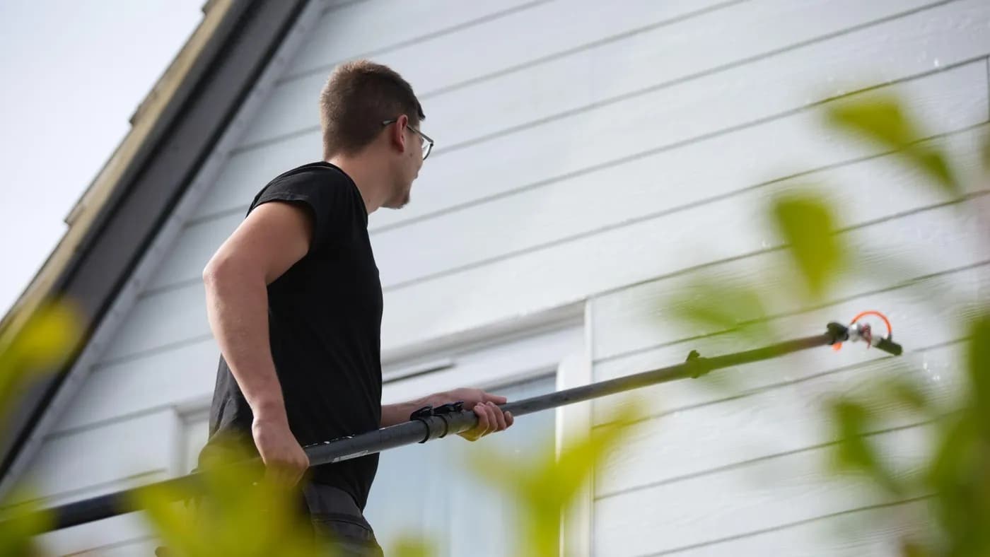 Operative holding a water-fed pole with a fan spray head against pale cladding, leaves blurred in the foreground