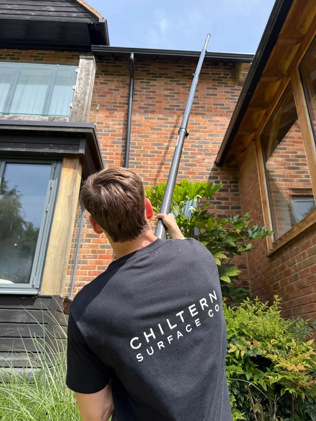 Operative seen from behind guiding a carbon gutter vacuum pole up to a black gutter on a two-storey brick and oak house