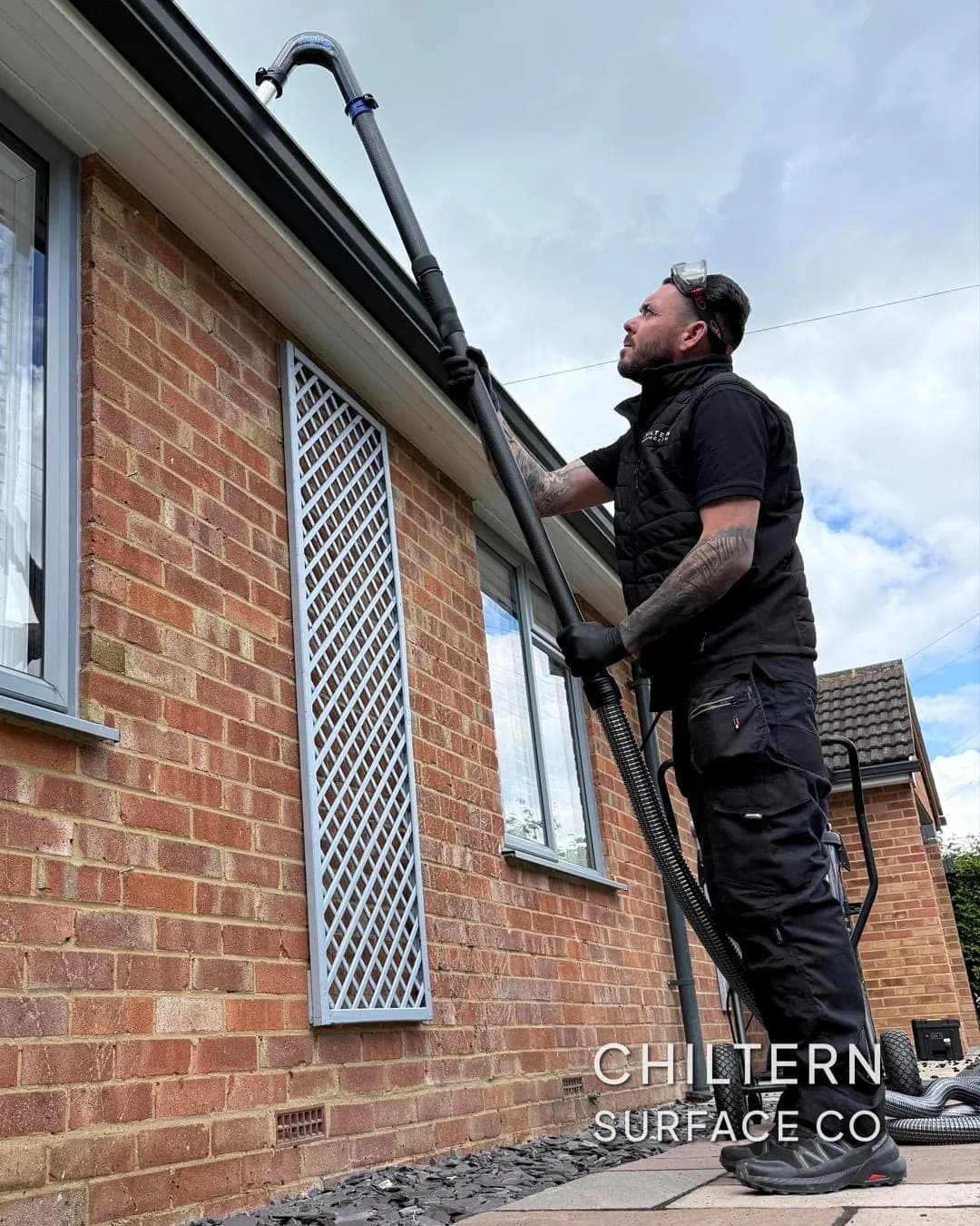Operative using a pole-fed gutter vacuum from a patio to clear the grey gutter of a red brick bungalow