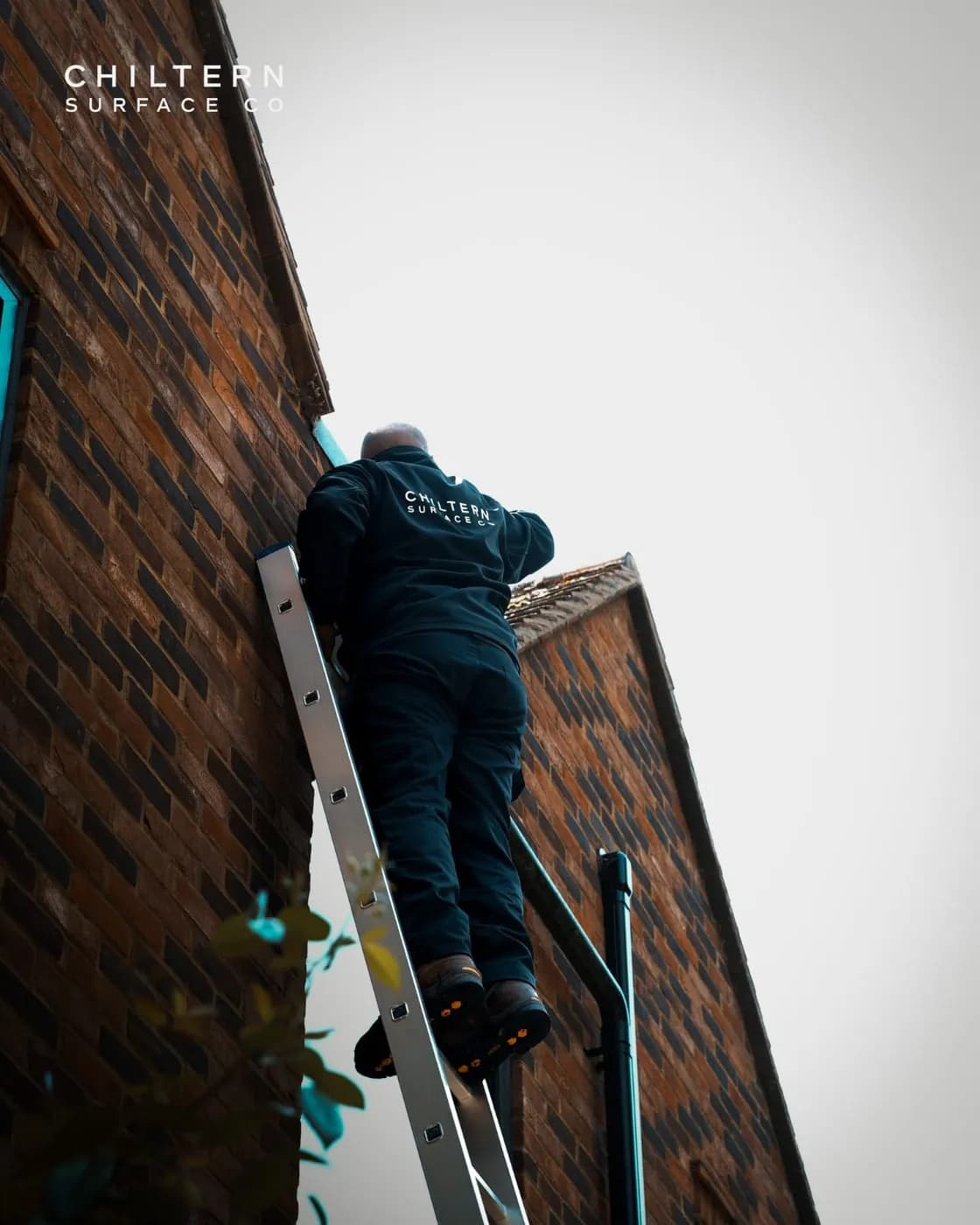 Operative in a branded jacket on an aluminium ladder at the gutter line of a brick house, a black downpipe beside him