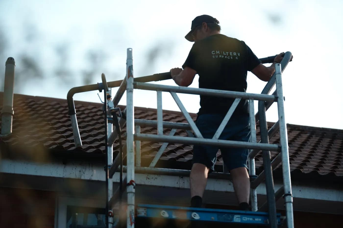 Operative on a mobile access tower gripping a gutter vacuum pole and hose above a stained white fascia and tiled roof