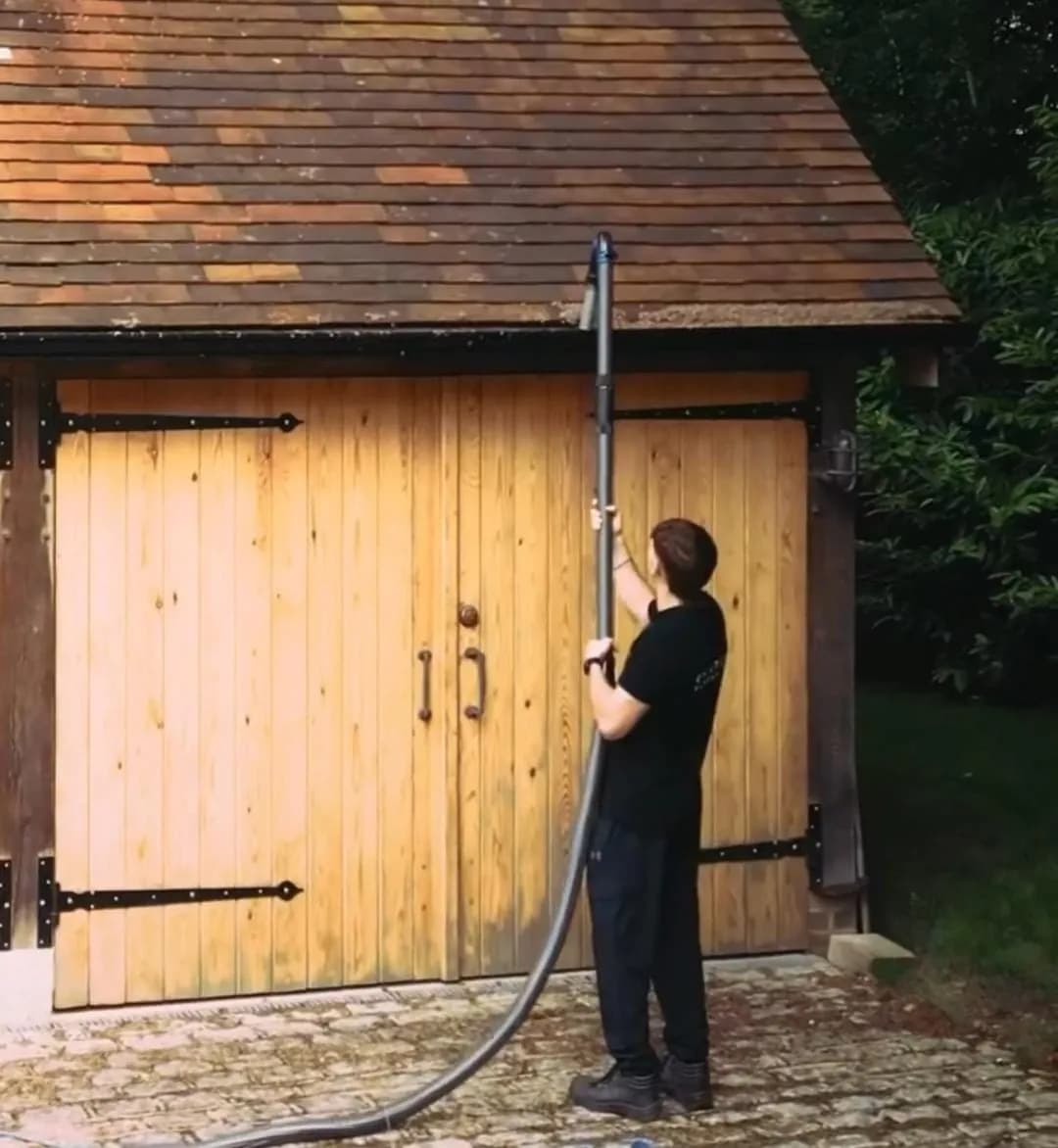 Operative using a gutter vacuum pole and hose at the eaves of a clay-tiled garage with oak doors and a setts drive