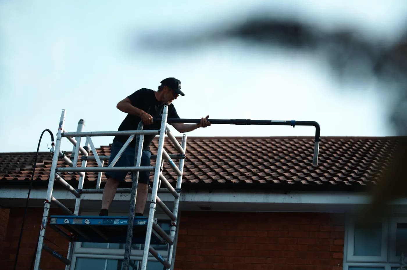 Operative on an aluminium access tower guiding a hooked vacuum nozzle over the gutter of a concrete-tiled roof
