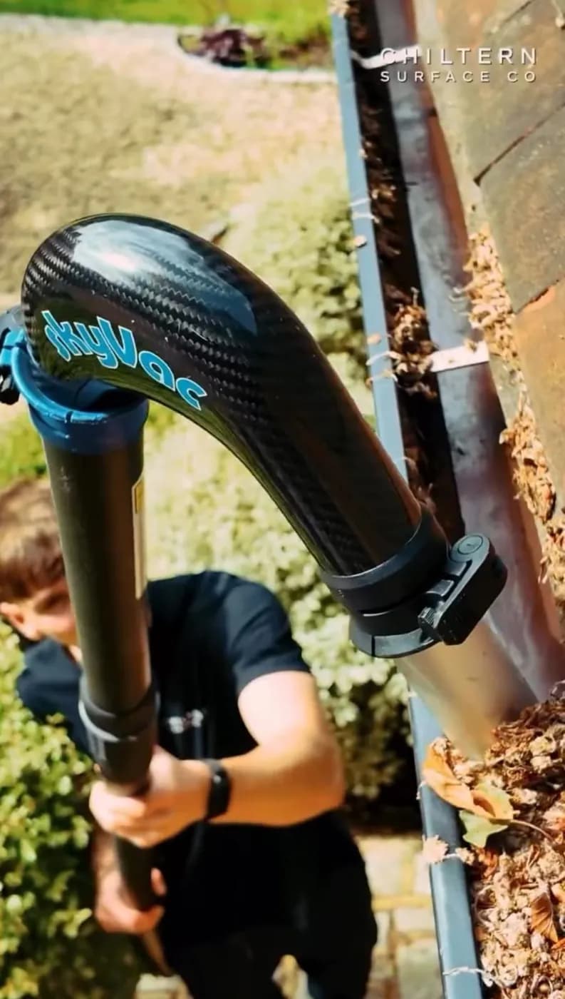 Curved carbon nozzle of a pole-fed gutter vacuum lowered into a grey gutter full of leaf litter, operative below
