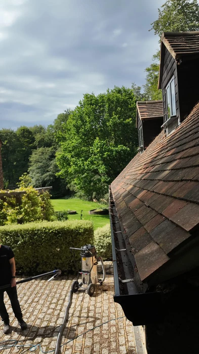 Clay-tiled roof with dormers and a black gutter holding leaf debris, a gutter vacuum and operative on setts below