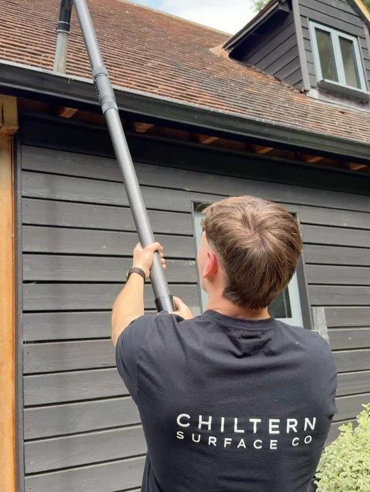 Operative in a branded T-shirt raising a carbon gutter vacuum pole to a black gutter below weathered clay roof tiles