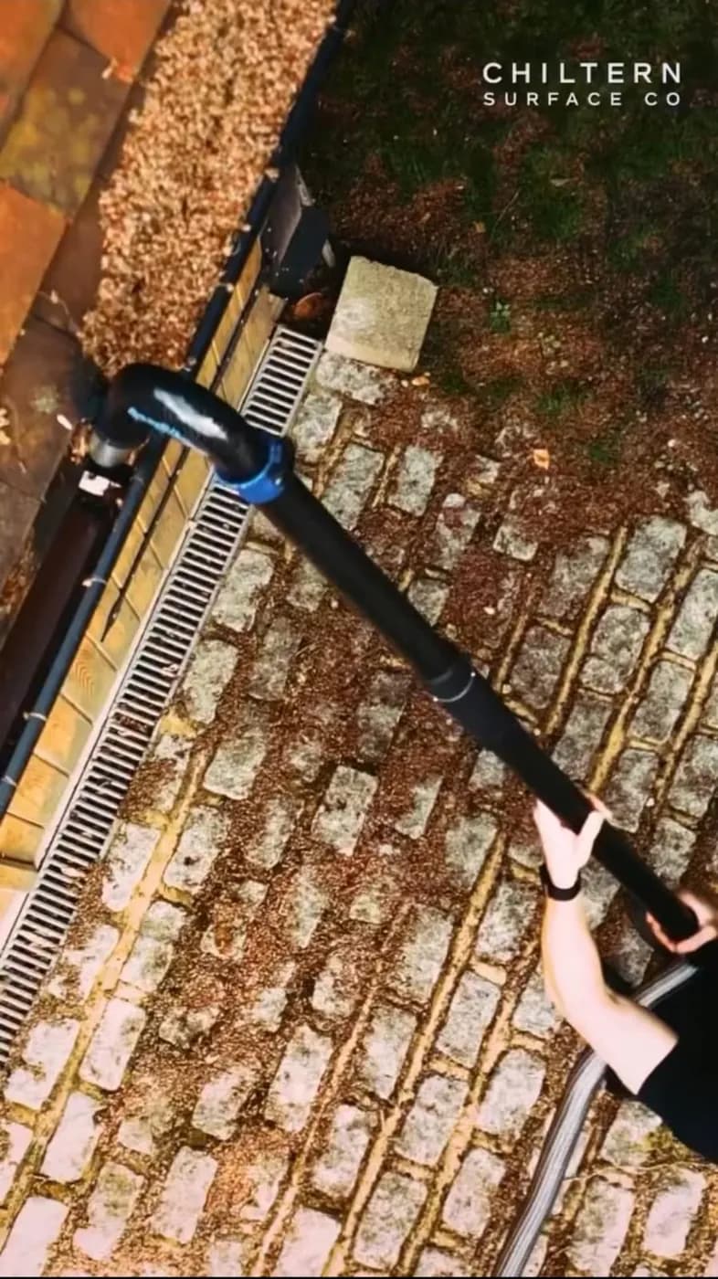 Looking down on a gutter vacuum pole held from the ground, its curved nozzle at a black gutter below leaf-strewn clay tiles
