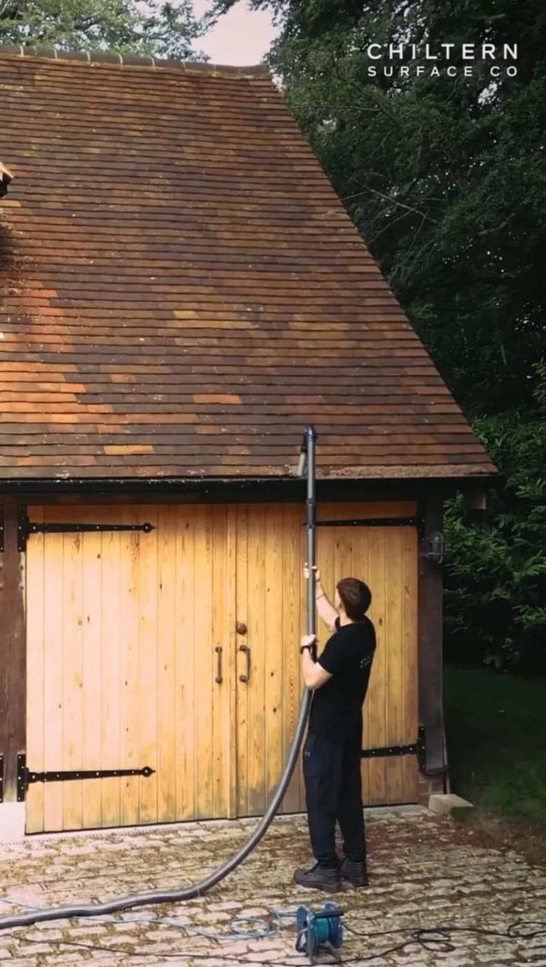 Operative holding a gutter vacuum pole up to the gutter of a clay-tiled oak garage, its hose trailing across cobbles