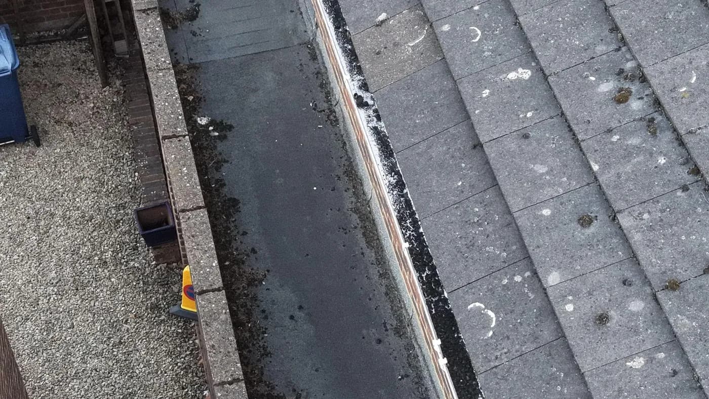 Overhead view of a lined box gutter behind a parapet, with moss clumps and debris beside a concrete-tiled roof