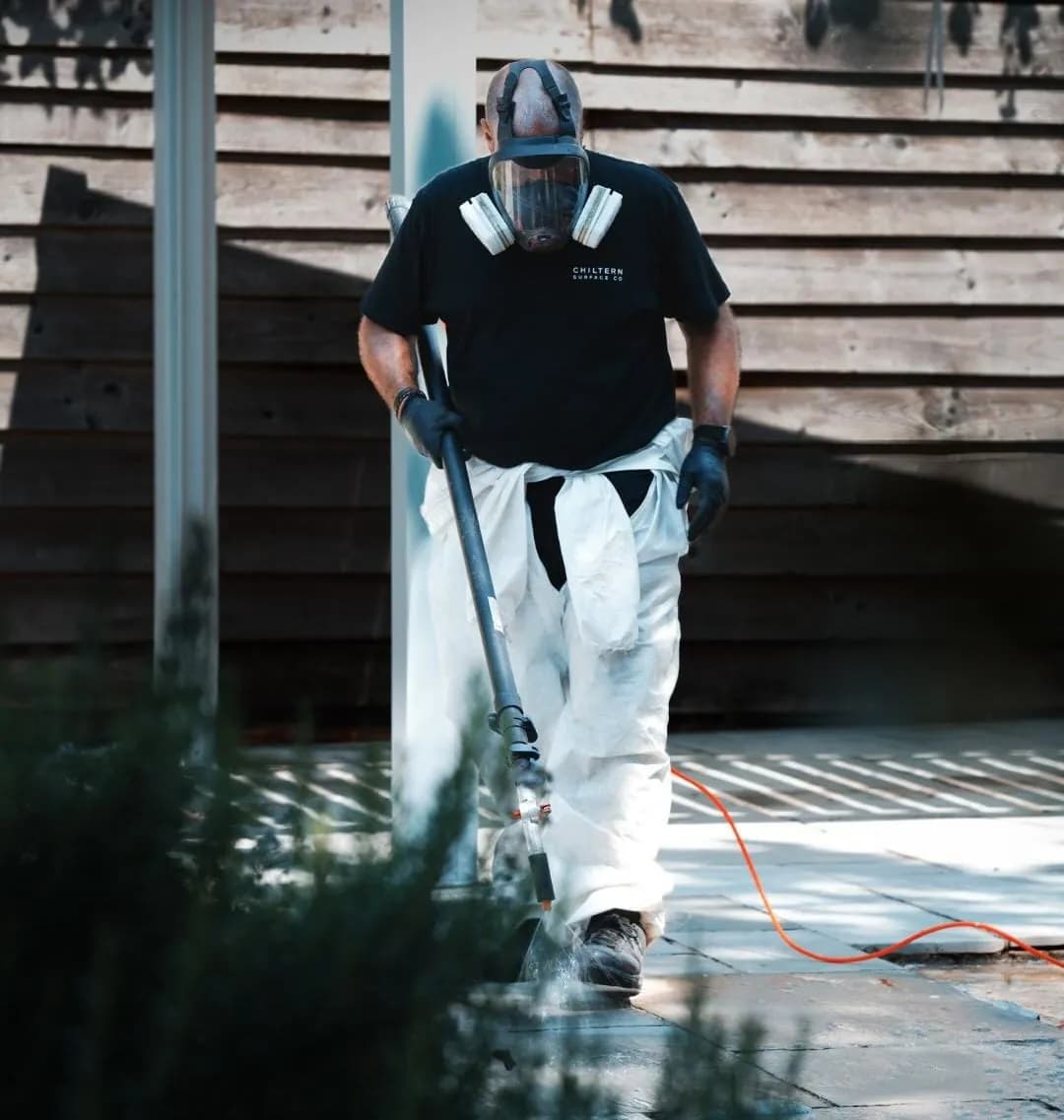 Operative in a respirator guiding a pole-mounted lance across pale stone paving beside timber cladding