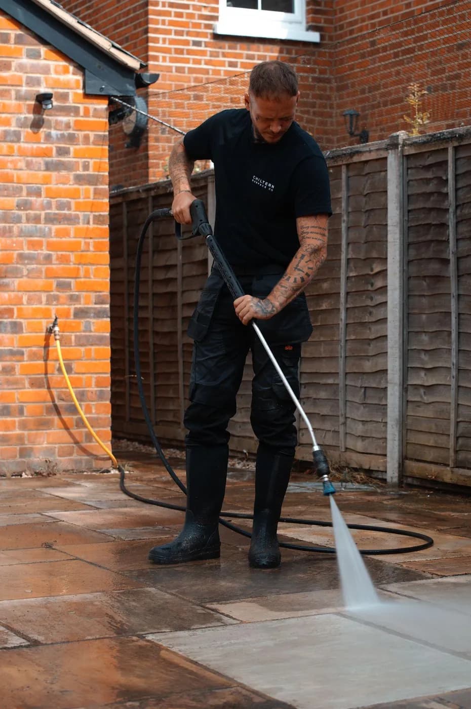 Operative in black workwear cleaning sandstone patio slabs with a lance, a clean pale strip showing against darker wet stone