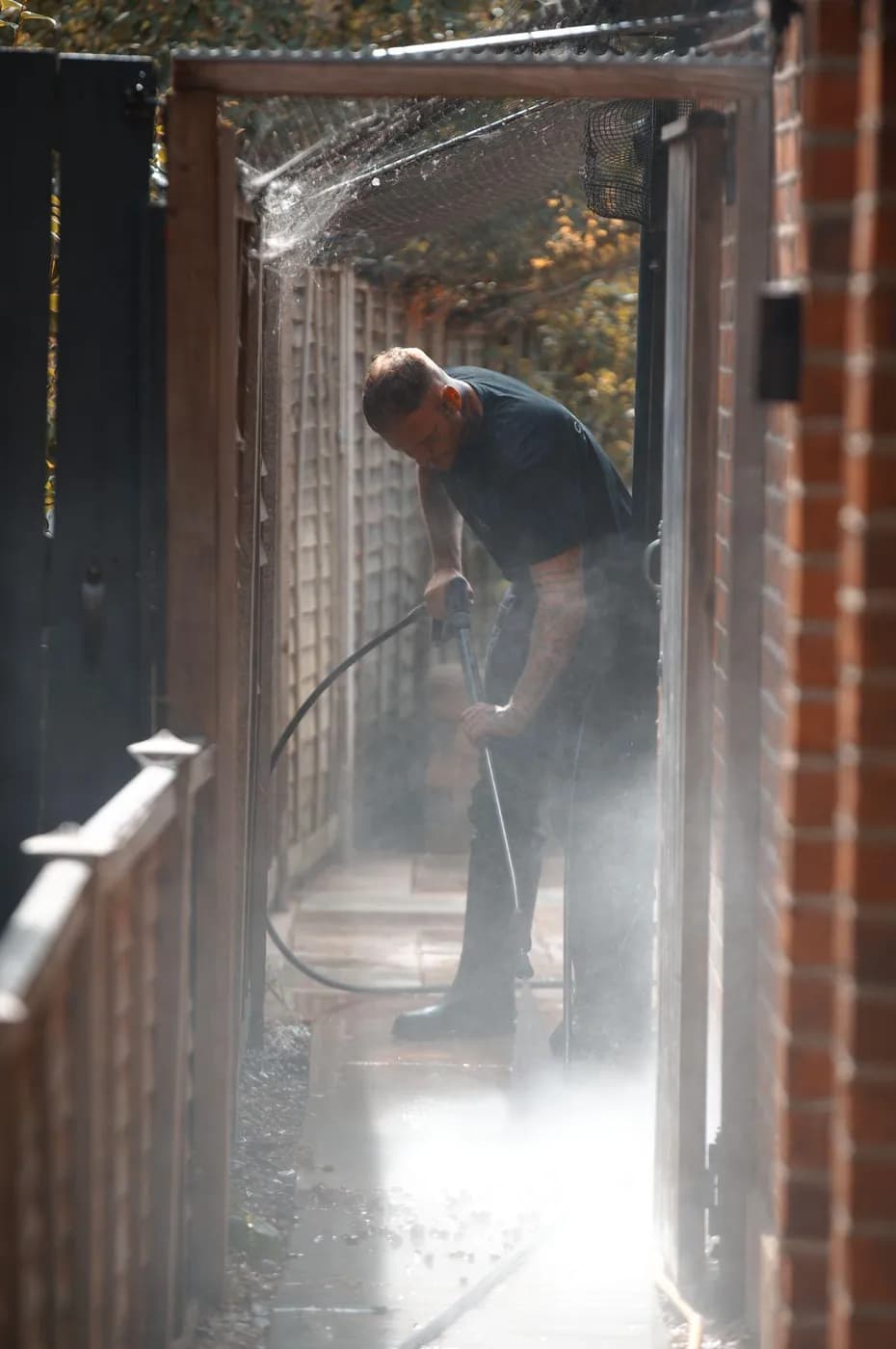 Operative cleaning a narrow paved side path between a timber fence and a brick wall, spray rising in the sunlight