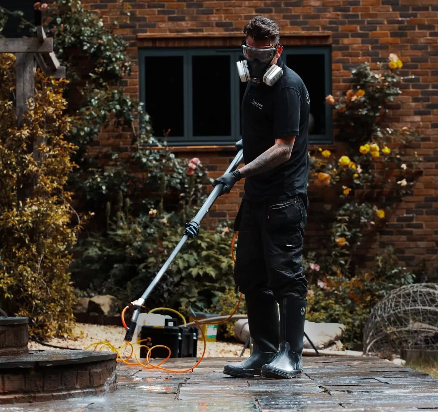 Operative in a respirator applying a low-pressure treatment by lance to stone paving beside a brick house