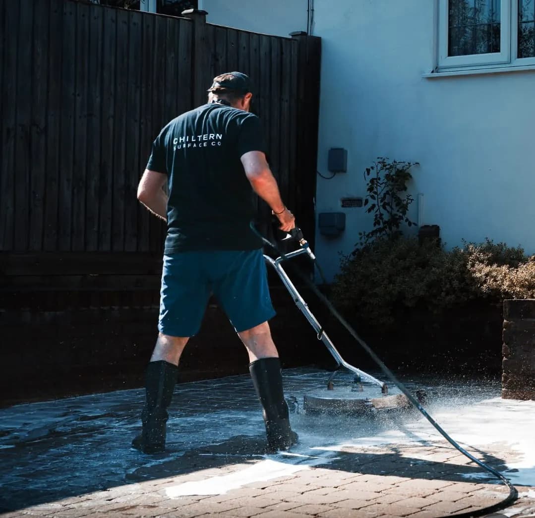 Operative guiding a rotary surface cleaner across wet block paving beside a rendered house, a timber fence and shrubs