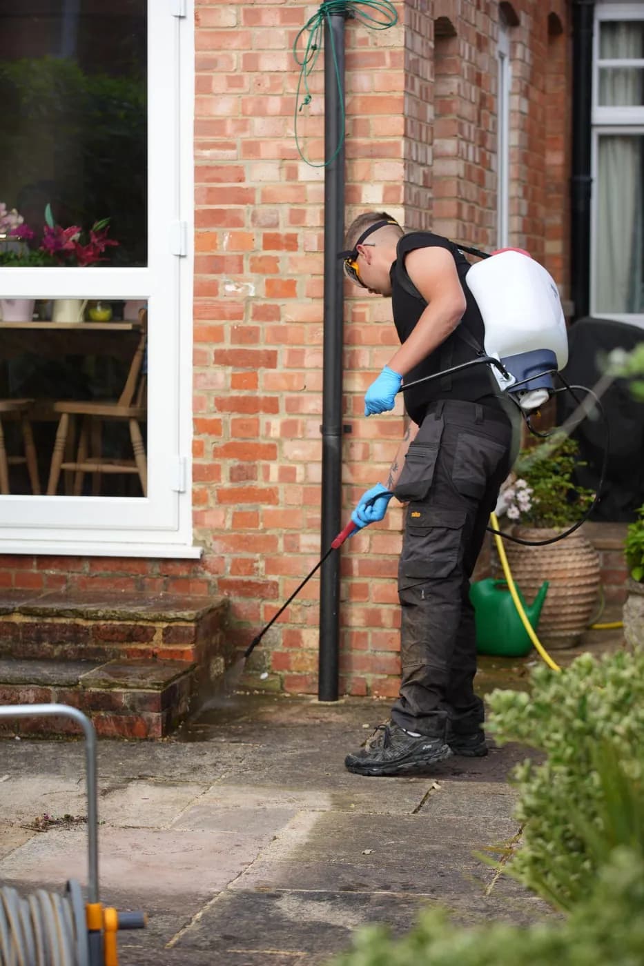 Operative with a knapsack sprayer applying treatment to sandstone paving at the foot of a red-brick wall beside garden steps