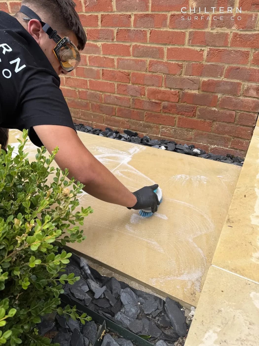 Operative in goggles and gloves hand-scrubbing a pale sawn sandstone step, with slate chippings and a box shrub alongside