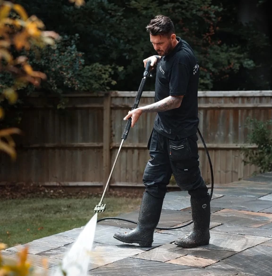 Operative cleaning dark Indian sandstone paving with a pressure lance in a fenced back garden beside a lawn
