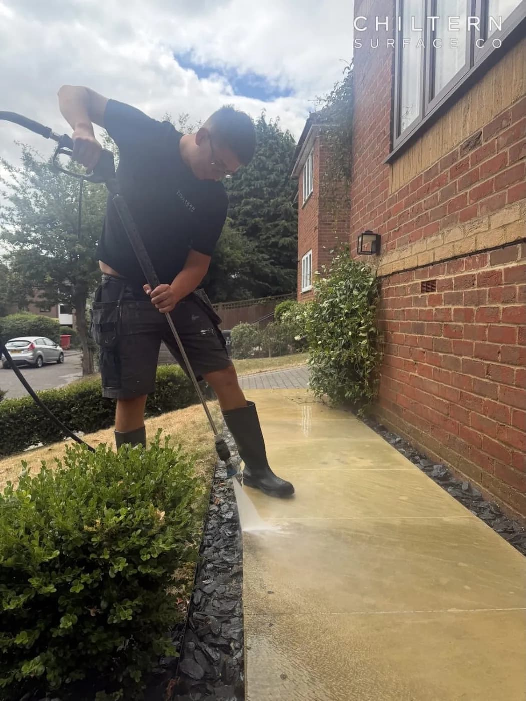 Technician using a pressure lance on a smooth buff sandstone path beside a brick house, with a box hedge and lawn alongside
